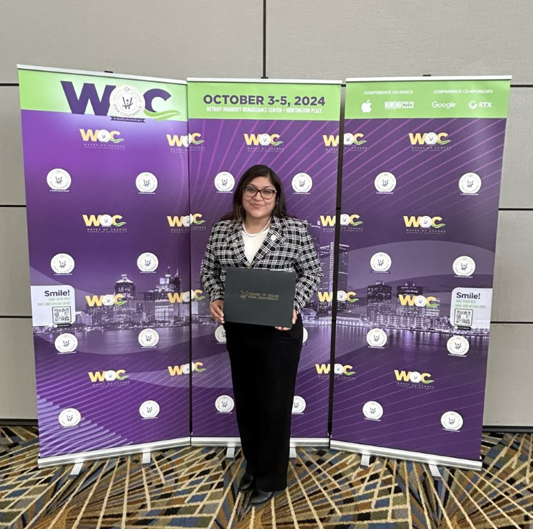 Michelle Dominguez A recipient of the Women of Color STEM Conference award is standing in front of the event's banner, holding a certificate in her hands. She is dressed in professional attire.