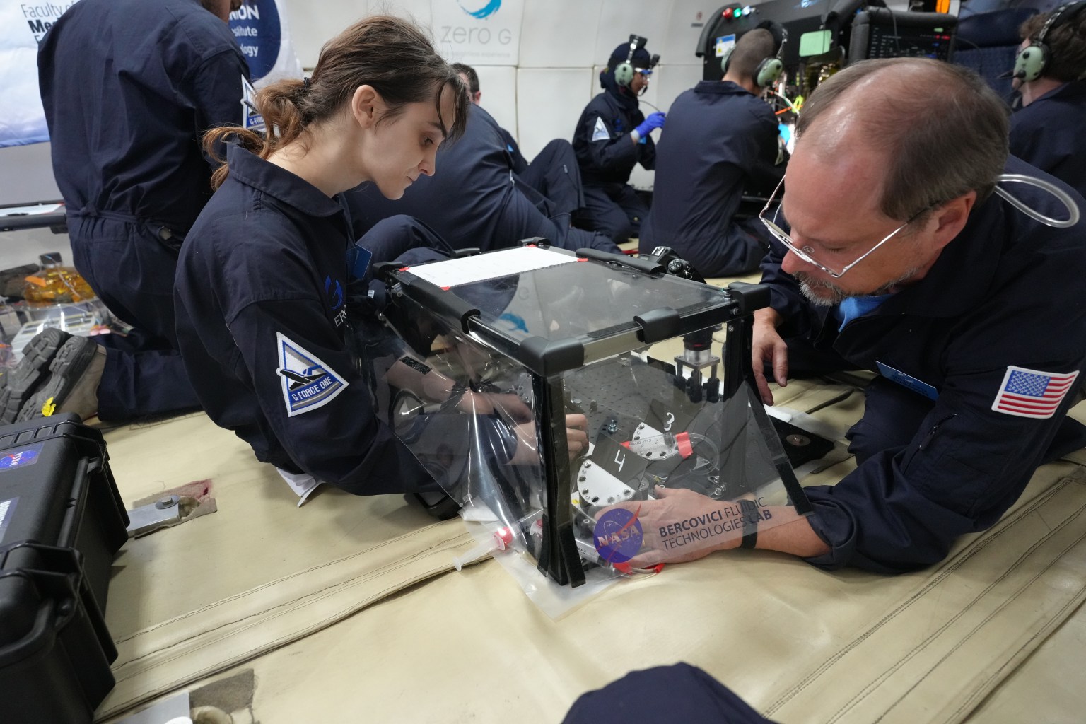 Two researchers sit on the floor of a parabolic jet making adjustments to their experiment housed in a clear frame. Other researchers are in the background.