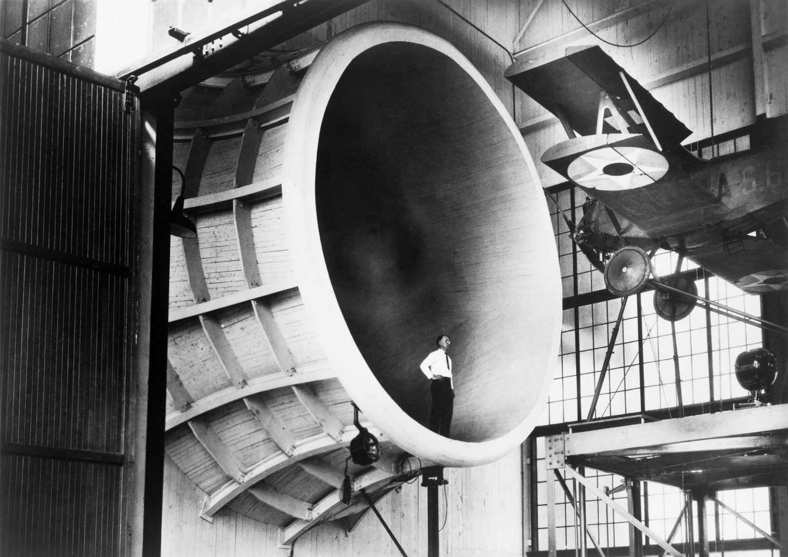 A man stands inside the entrance of a tunnel, looking up at a small propeller plane. The tunnel, viewed from the side, is like a cylinder with sides curving inward. The photo is in black and white.