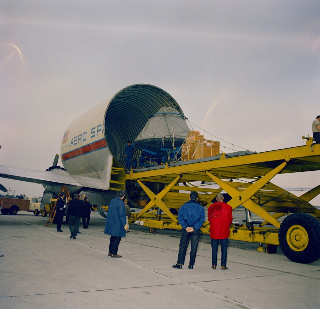 Image of workers offloading a .conical spacecraft from an open cargo plane.