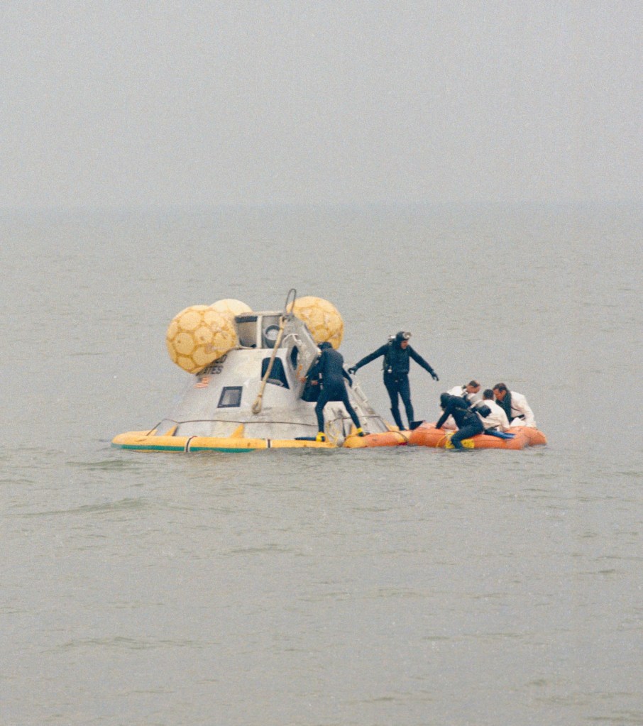 A conical spacecraft floating on water with several men in life rafts around it.