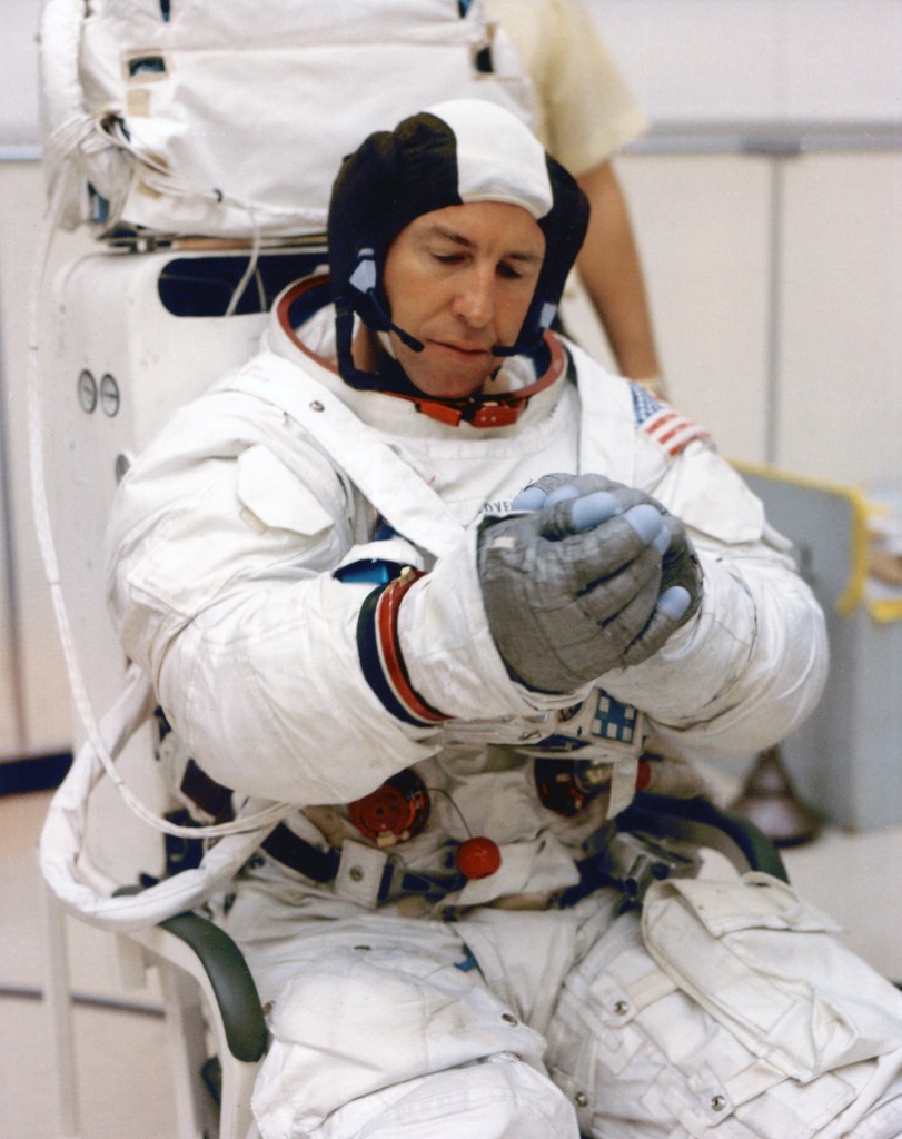 A man wearing a white spacesuit and black and white communications cap is putting on his gloves.