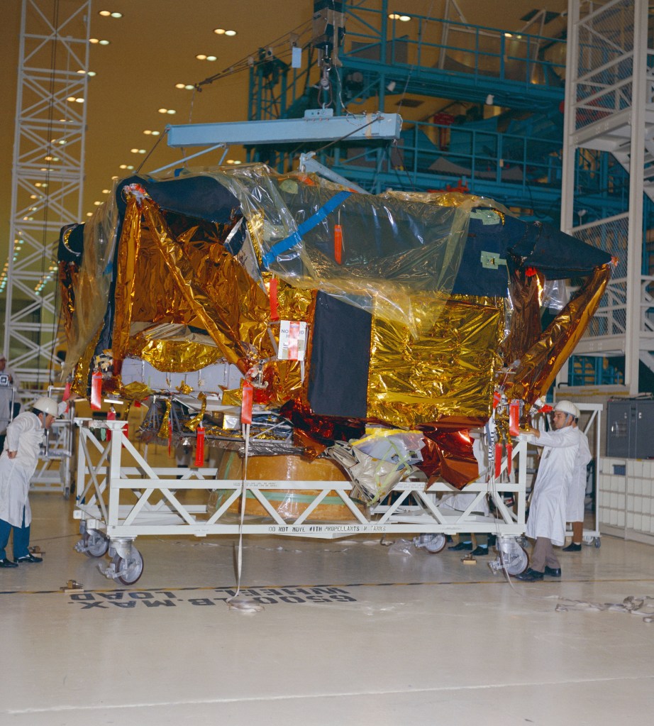 A gold foil covered rectangular looking spacecraft sits on a dolly in a high bay.