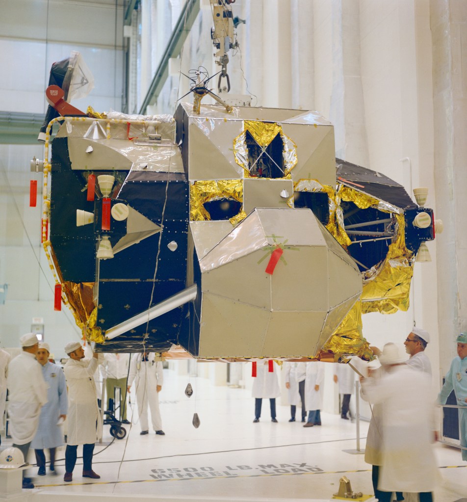 Image of an irregularly-shaped spacecraft, a Lunar Module, being lifted in a high bay.