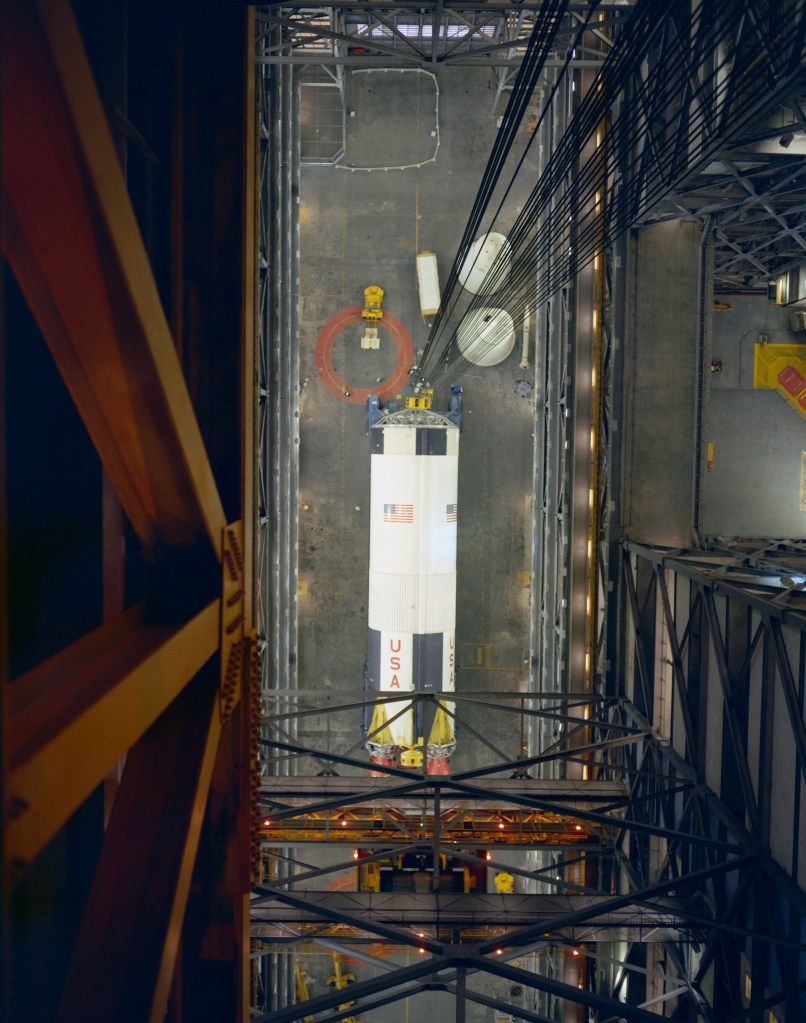 A large white and black cylindrical rocket stage sits on the floor of a large assembly building