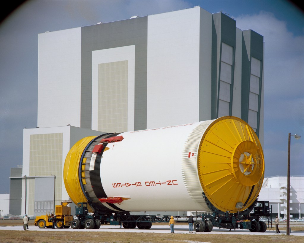 A large cylindrical white rocket stage lying on its side outside a large white and black assembly building