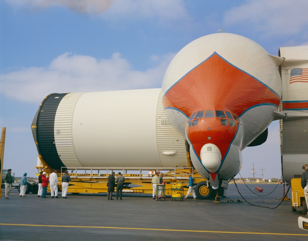 A large mostly white cylindrical rocket stage is being offloaded from an open cargo plane.