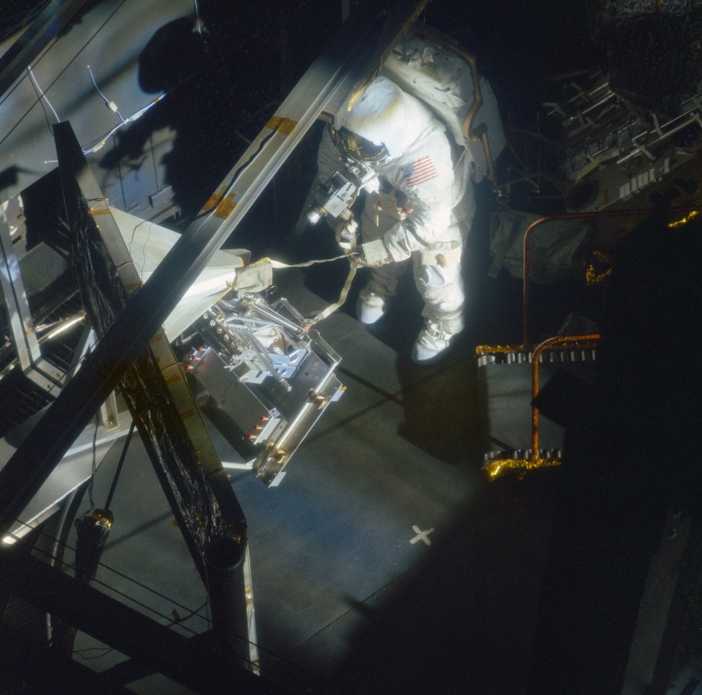 An astronaut clad in a white spacesuit works with experiment hardware inside a vacuum chamber