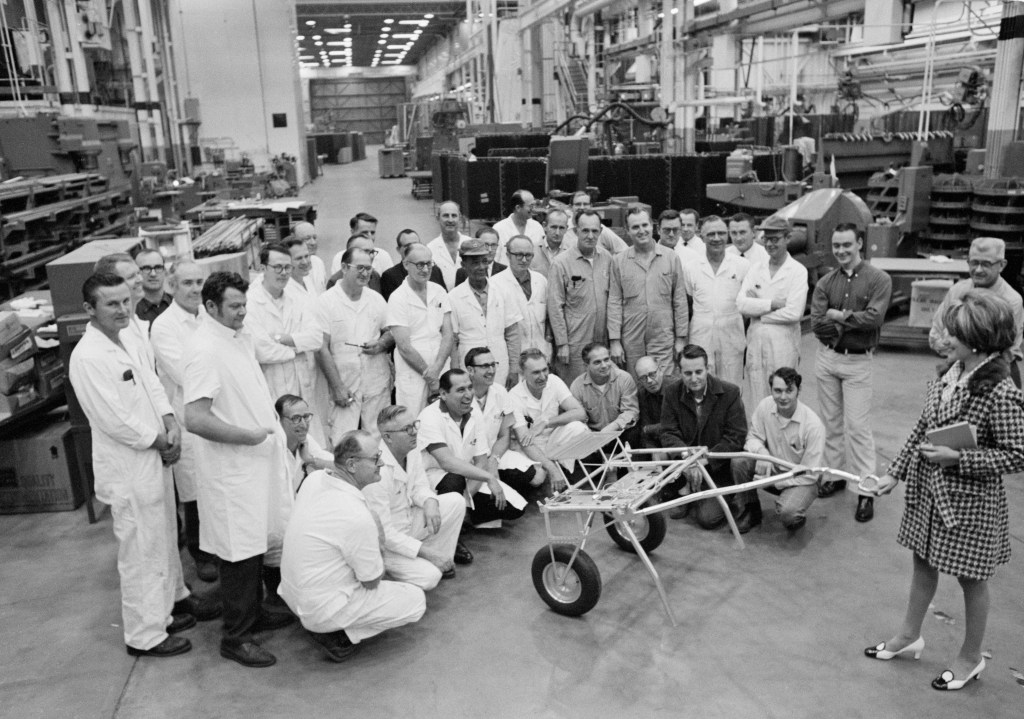 Black and white image of a group of men standing facing a woman holding a golf cart like vehicle.