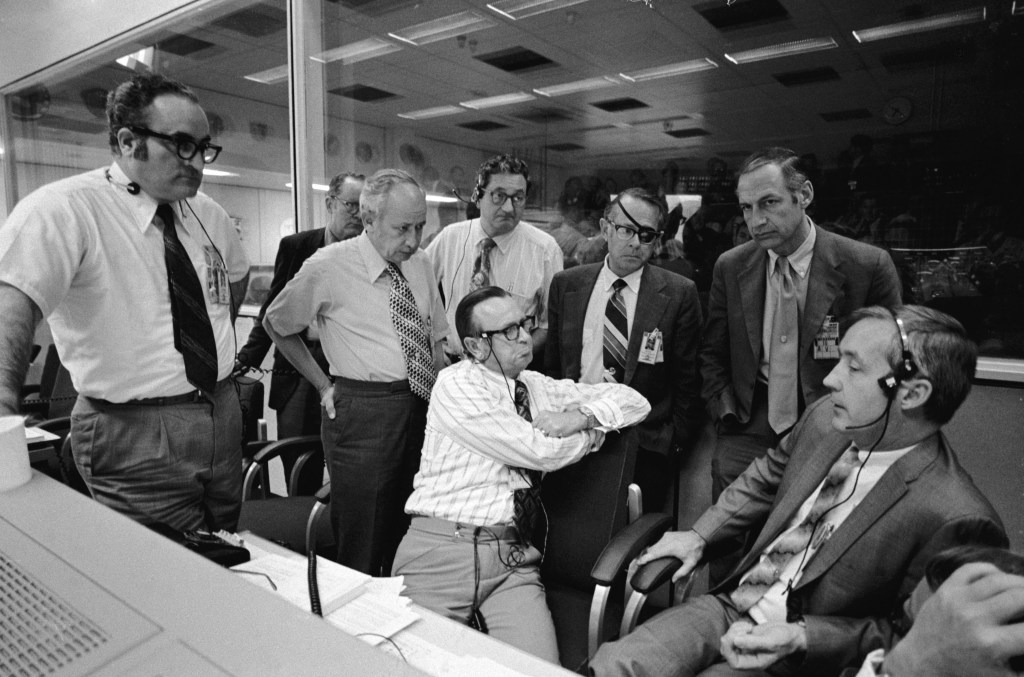 Black and white image of a group of serious looking men, discussing an engineering problem in Mission Control.