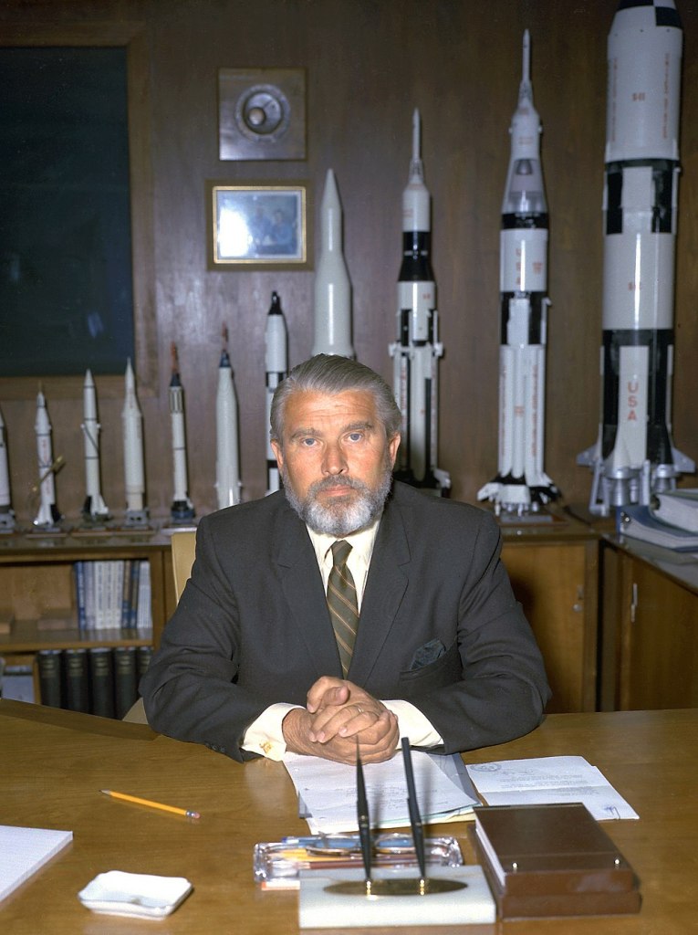 A bearded middle aged man in a business suit sitting at a wooden desk with a series of model rockets behind him.