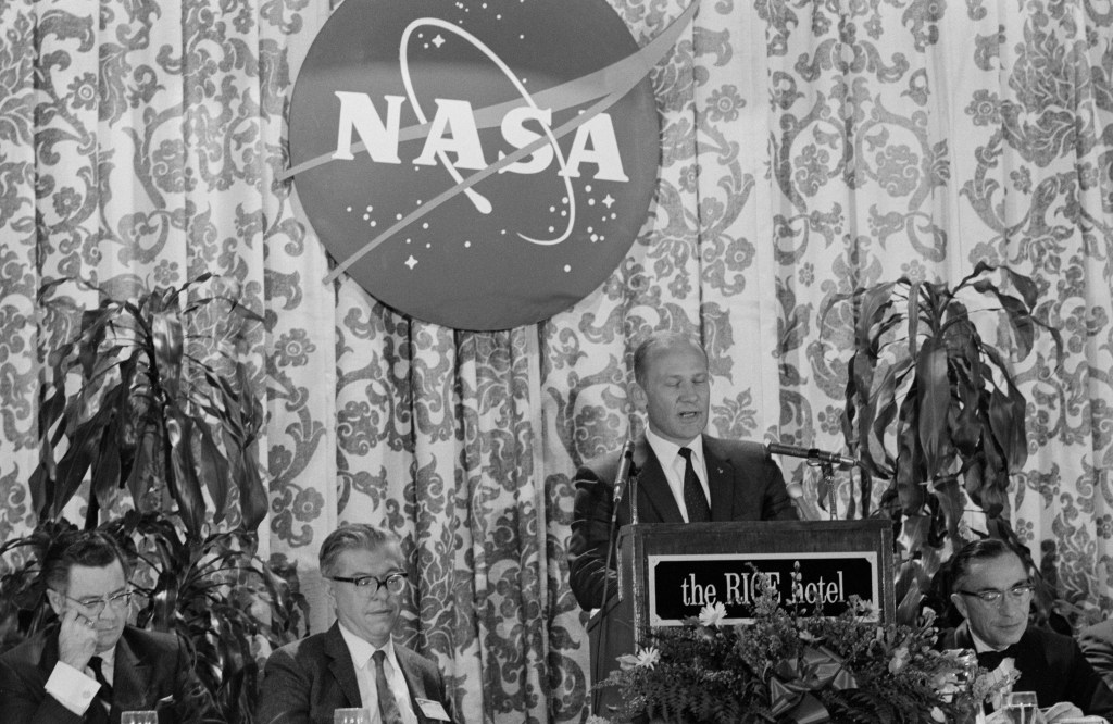 Black and white image of a man - an astronaut - standing at a podium, speaking to an off-camera crowd, with a large NASA logo behind him.