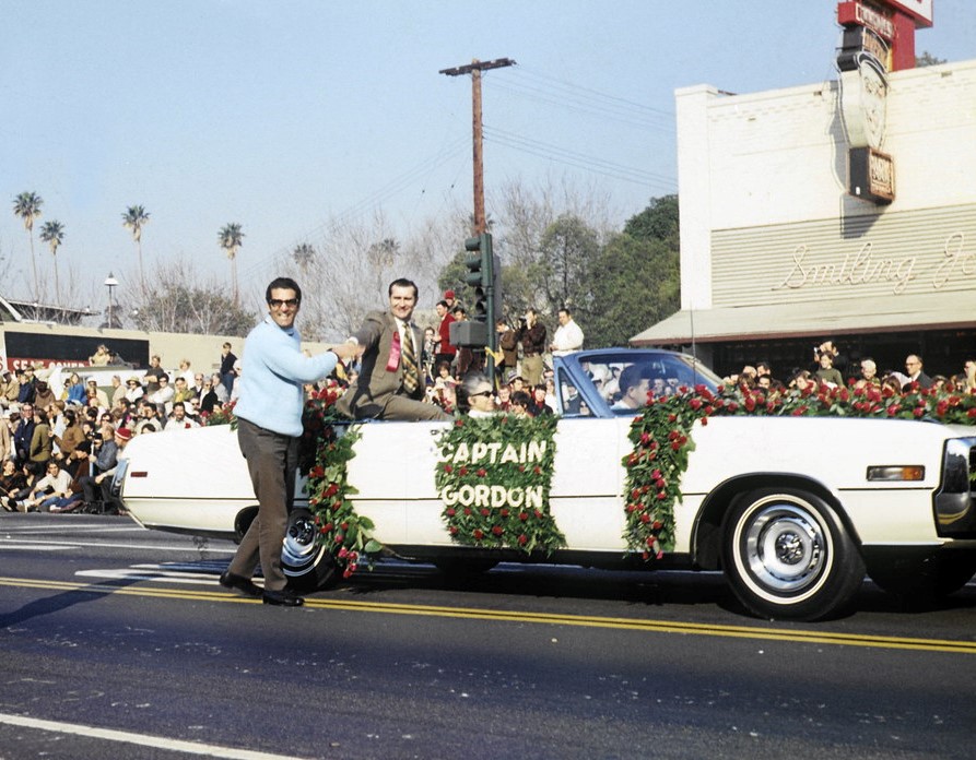 A white convertible limousine in a parade, with a sign saying 