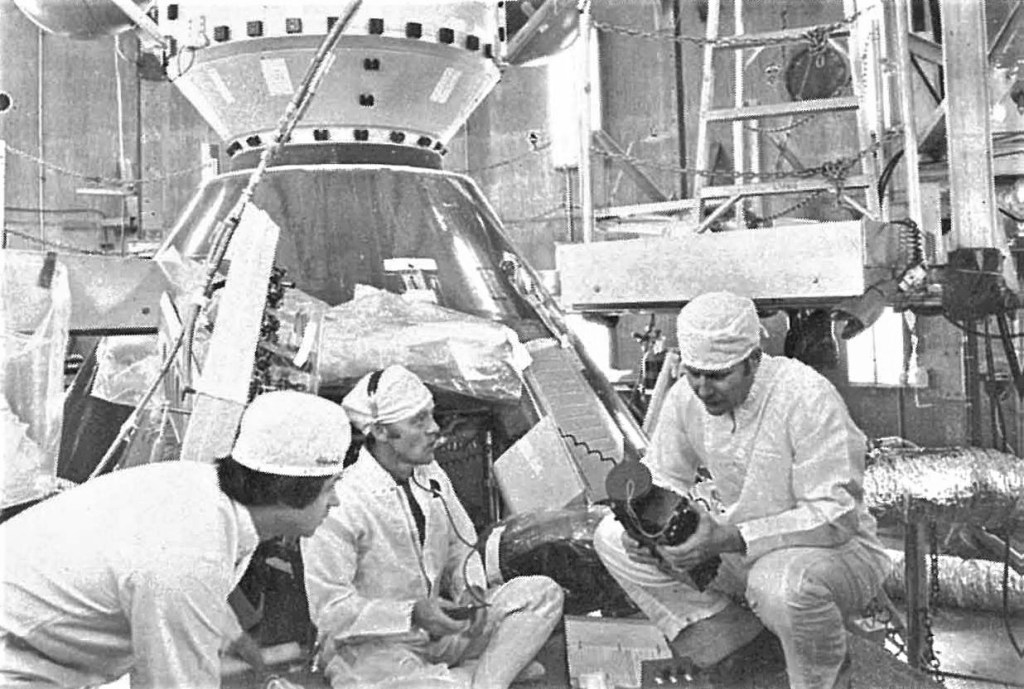 Black and white image of several white coated technicians with a partial view of a spacecraft in the background