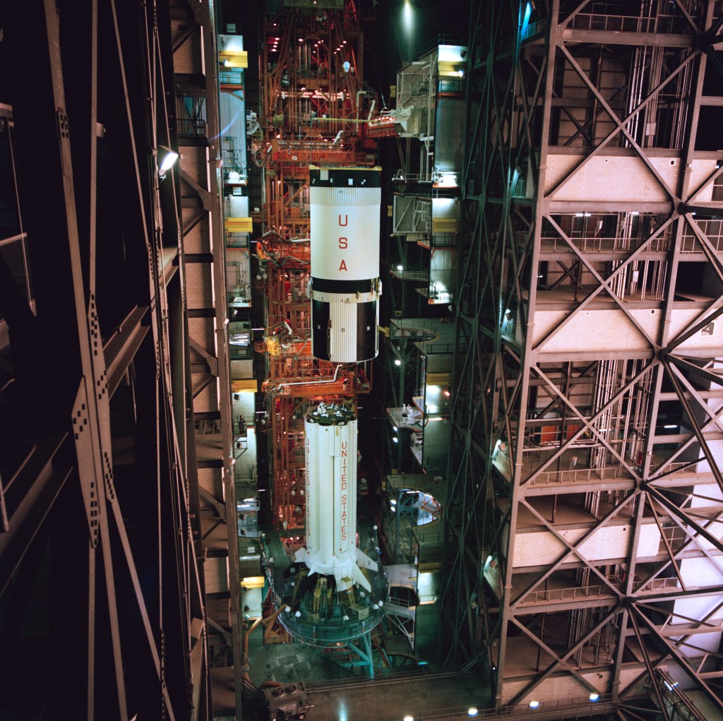 A rocket's white and black second stage is lowered onto its white first stage inside a large assembly building