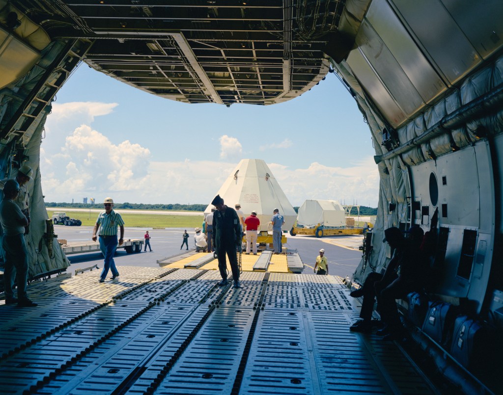 A view from inside a cargo aircraft through the open aft cargo door on a runway, with two white spacecraft being unloaded