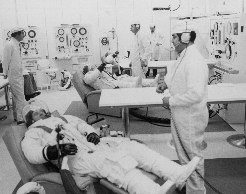 Black and white image of three astronauts in recliners wearing white spacesuits with technicians assisting them