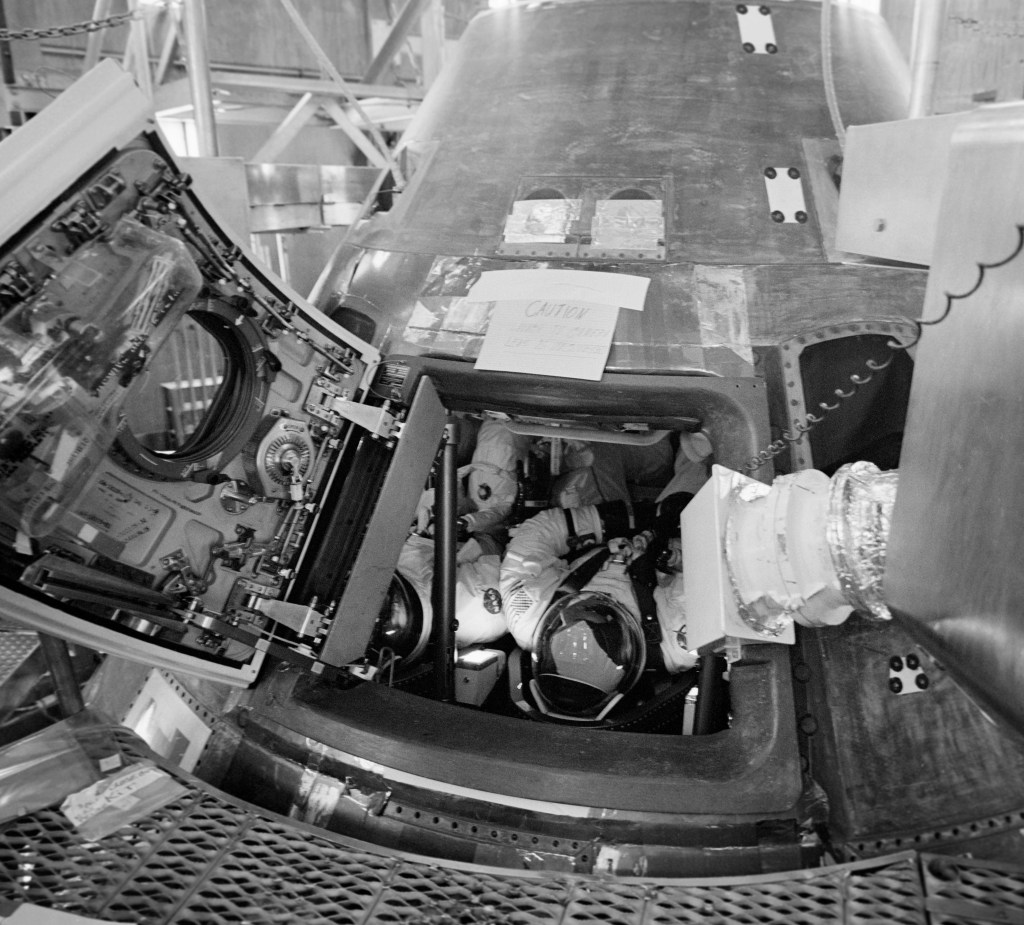 Black and white image of a cone-shaped spacecraft, with three spacesuited astronauts visible through the open hatch