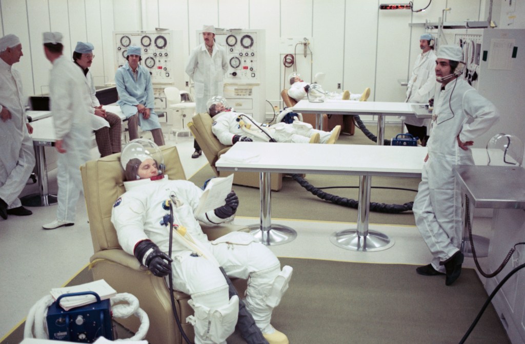 Color image of three astronauts in white spacesuits in recliners with technicians assisting them