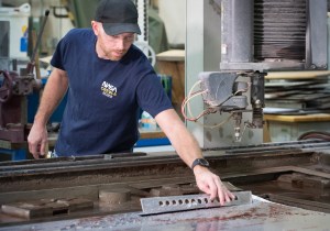 A man wearing a navy-blue shirt, a black baseball cap, and transparent safety glasses leans over a table while holding a strip of material that was cut from a water jet machine.