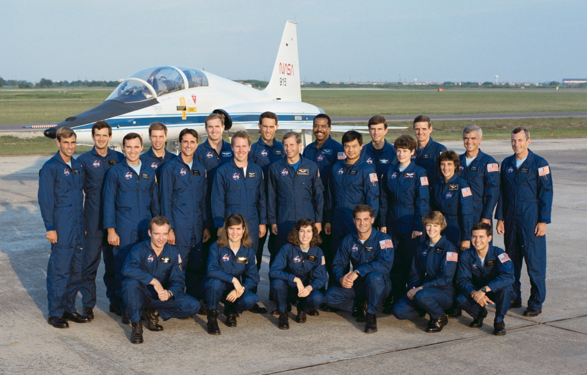 Outdoor group photo of 23 men and women wearing blue overall flight suits, with a jet airplane in the background.