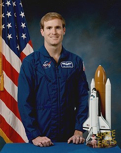 Portrait of a man wearing a blue jumpsuit with a partially visible US flag in the background.