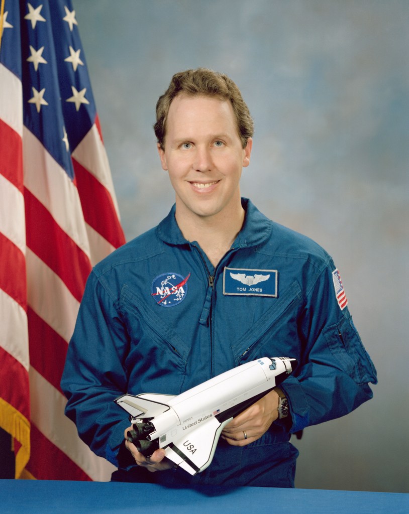 Portrait of a man wearing a blue jumpsuit with a partially visible US flag in the background.