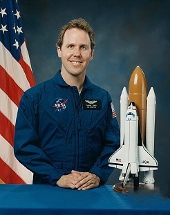 Portrait of a man wearing a blue jumpsuit with a partially visible US flag in the background.