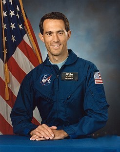 Portrait of a man wearing a blue jumpsuit with a partially visible US flag in the background.