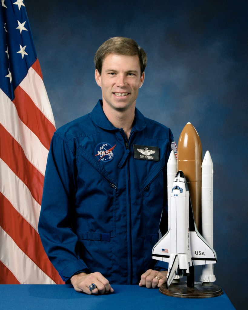 Portrait of a man wearing a blue jumpsuit with a partially visible US flag in the background.