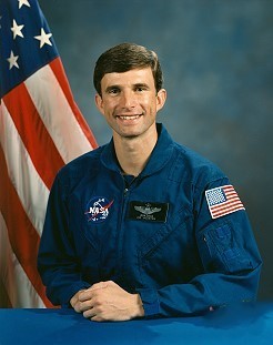 Portrait of a man wearing a blue jumpsuit with a partially visible US flag in the background.