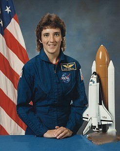 Portrait of a woman wearing a blue jumpsuit with a partially visible US flag in the background.