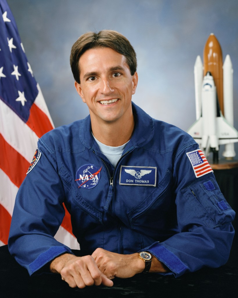 Portrait of a man wearing a blue jumpsuit with a partially visible US flag in the background.