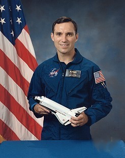 Portrait of a man wearing a blue jumpsuit with a partially visible US flag in the background.