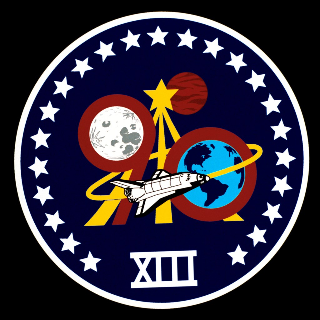 A round patch with a dark blue background, stylized images of the Moon, the Earth and a space shuttle superimposed on the astronaut symbol, with the Roman numeral XIII at the bottom and 23 white stars around the edge