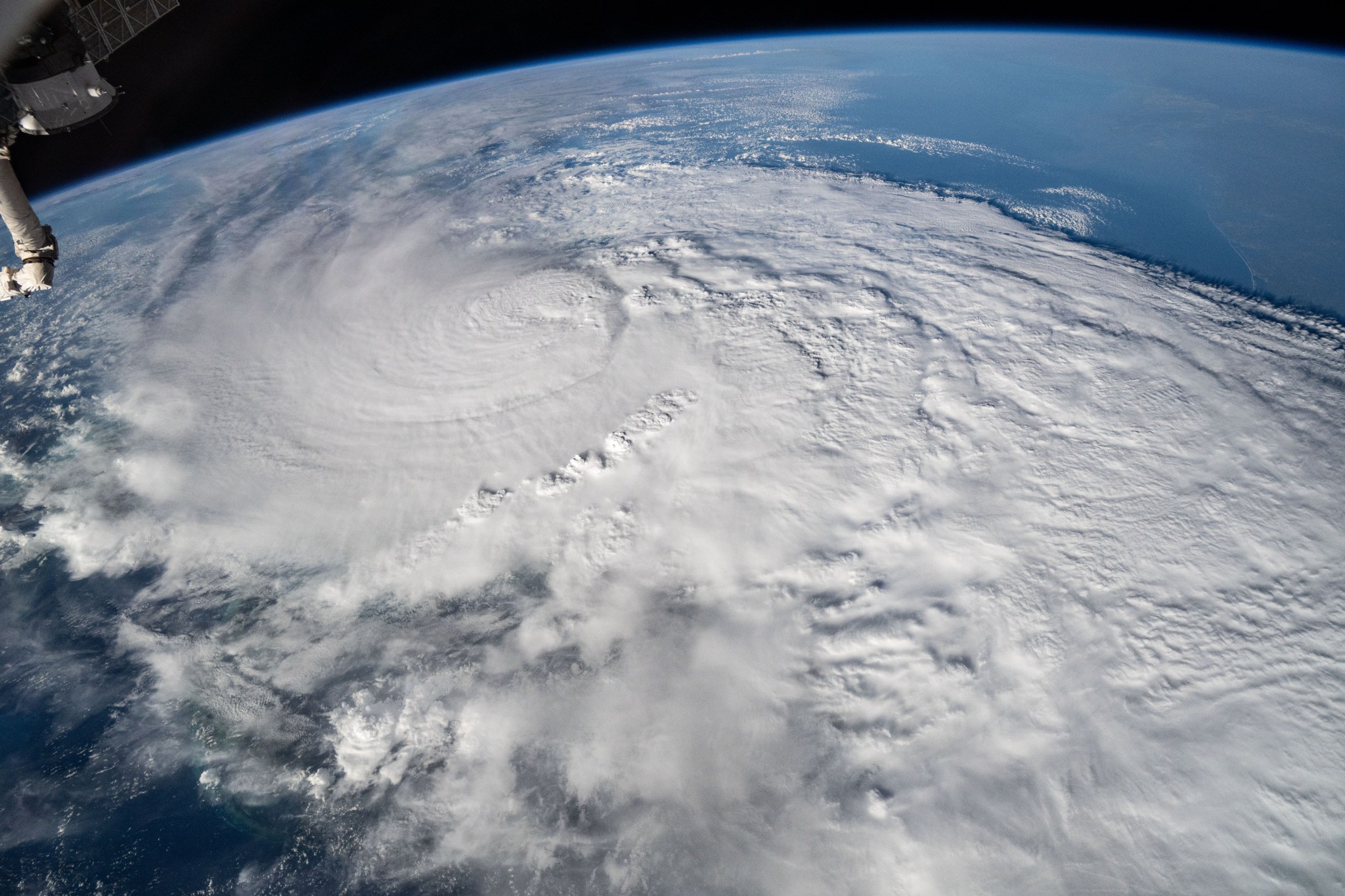 Milton is a large white spiral and a long cloud trail extending across this image. A portion of the space station is visible in the upper left corner, with blue ocean on Earth in the upper right.