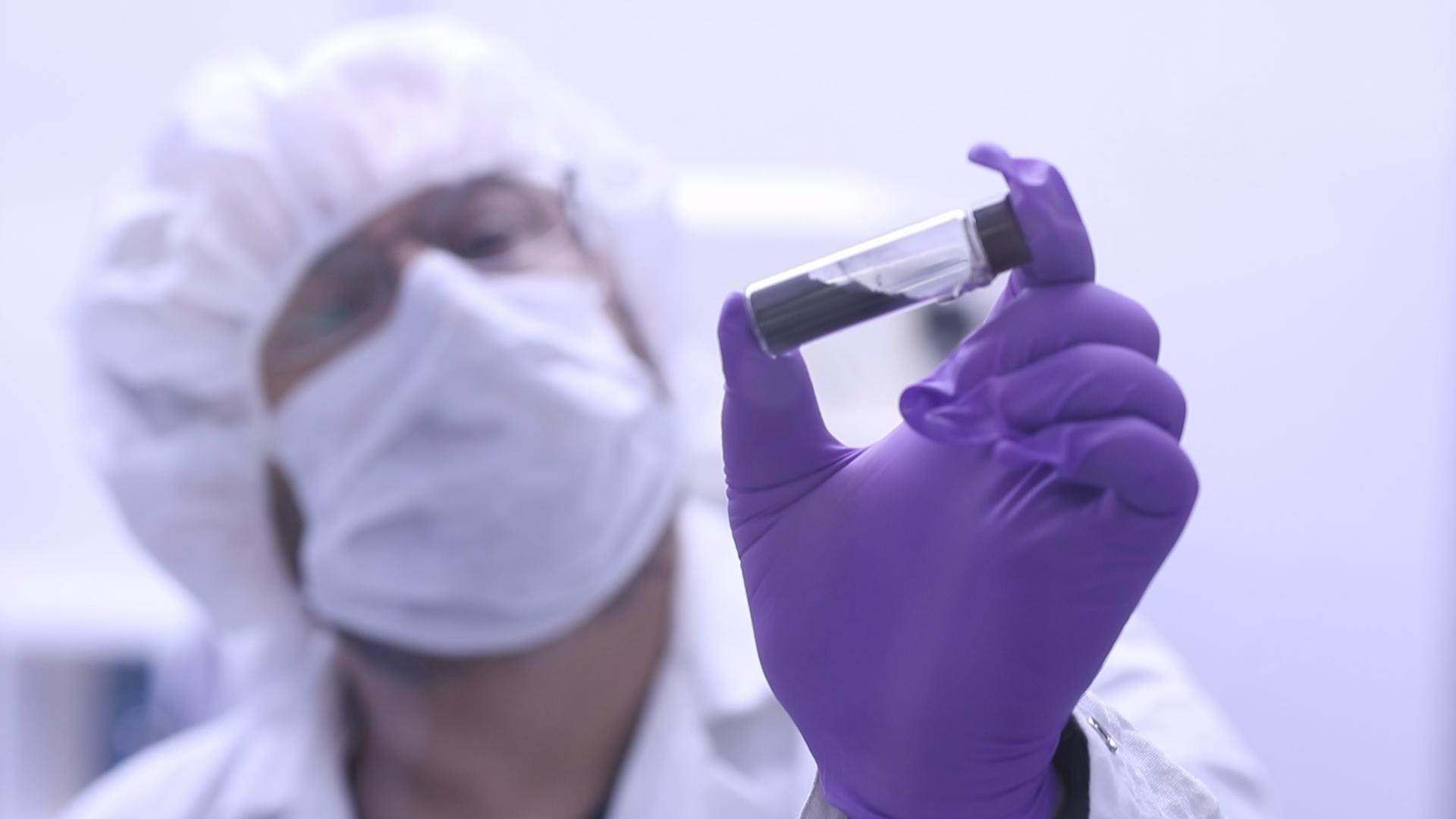 A scientist in a white coat, mask, hair net, and purple gloves holds up a clear vial with a black substance inside near the camera.