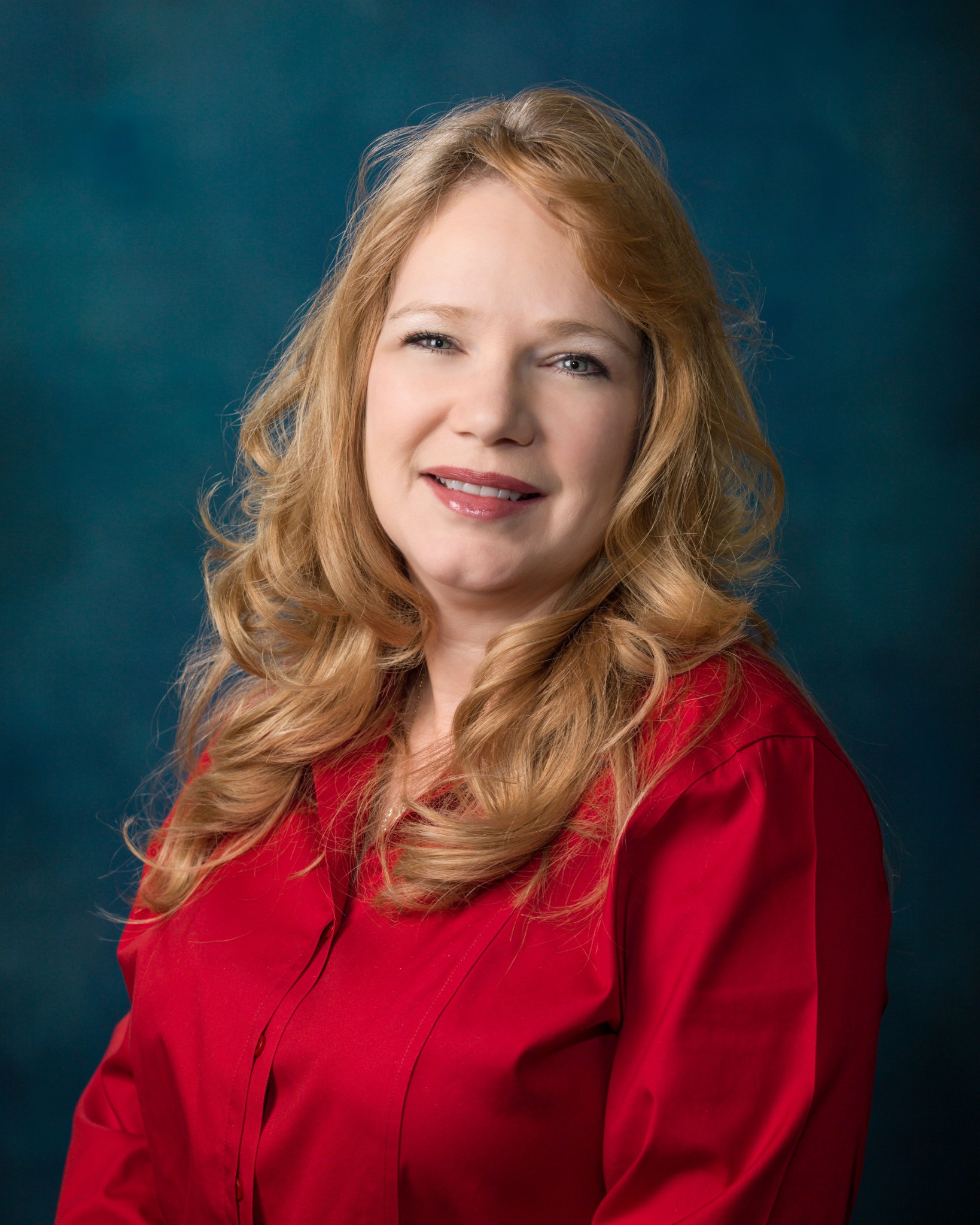 A red-haired woman in a red shirt smiles as she looks at the camera