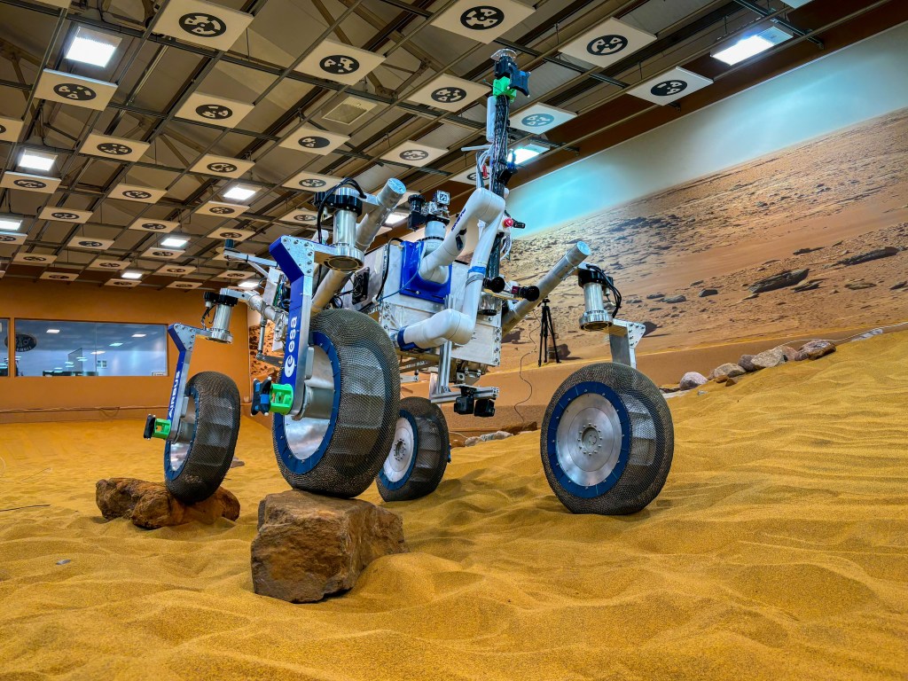 A white and blue test rover on sandy red Martian-simulated terrain traverses over large boulders, testing shape memory alloy spring tires.