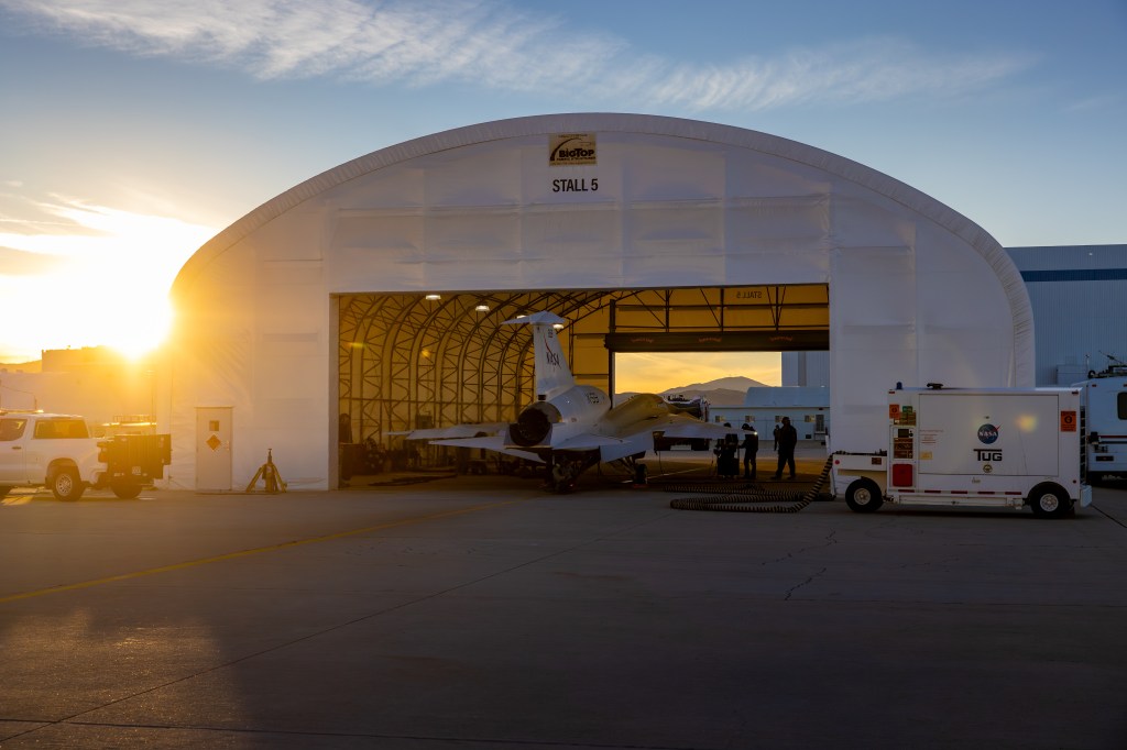 NASA’s X-59 sits partially outside a white hangar at Lockheed Martin’s Skunk Works Facility in Palmdale, California, during sunset, as engineers prepare it for afterburner testing.