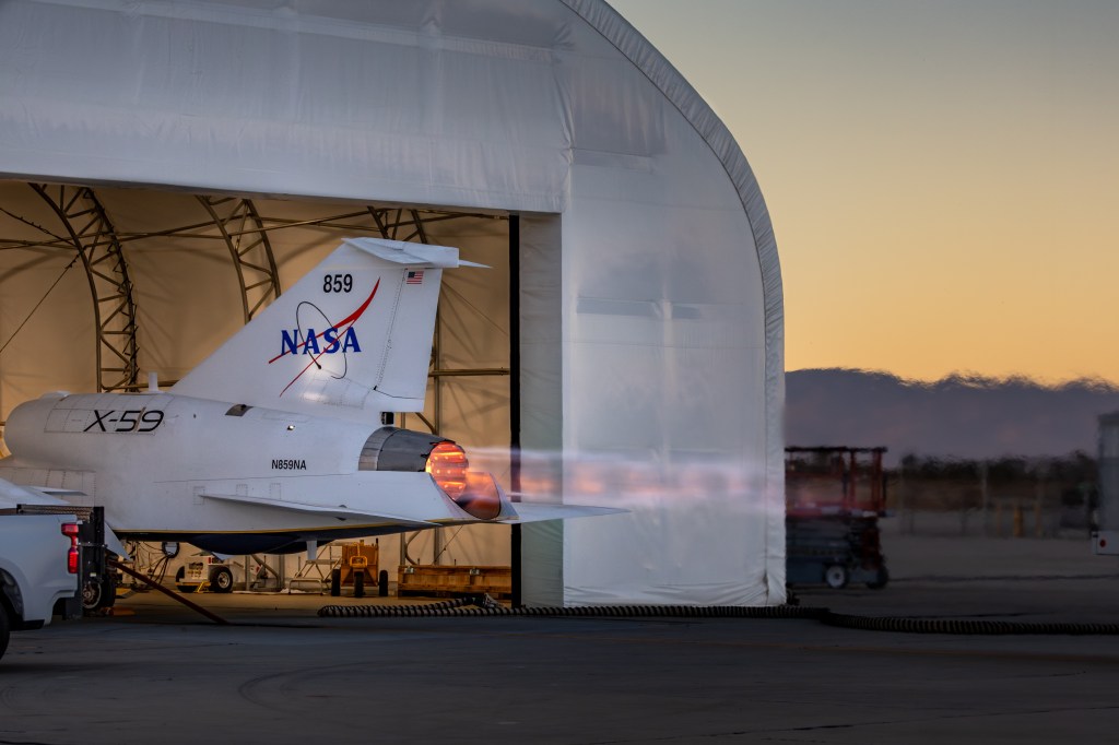 NASA’s X-59 sits partially in a white hangar and emits bright flames with visible shock diamonds from its engine against an evening sky during maximum afterburner testing outside Lockheed Martin’s Skunk Works Facility in Palmdale, California.