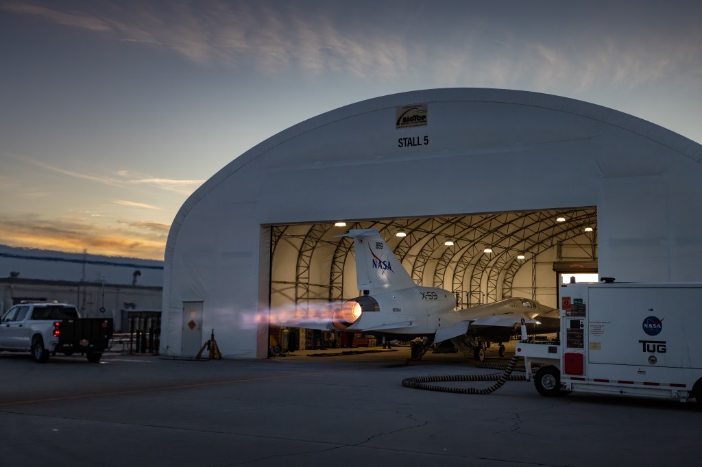 NASA’s X-59 sits partially in a white hangar and emits bright flames with visible shock diamonds from its engine against a darkening sky during maximum afterburner testing outside Lockheed Martin’s Skunk Works Facility in Palmdale, California.