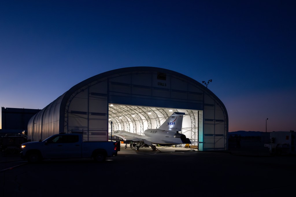 NASA’s X-59 sits partially outside a white hangar at Lockheed Martin’s Skunk Works Facility in Palmdale, California after sunset.