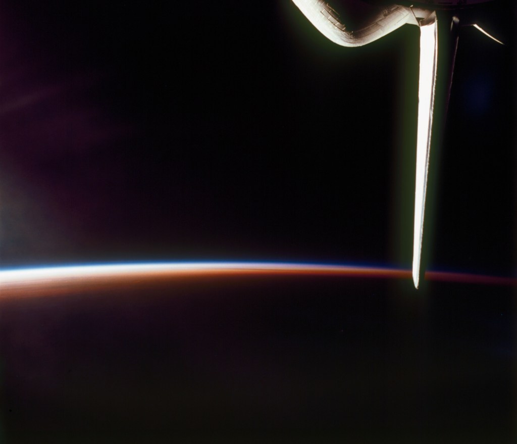 Mostly black image of a thin crescent of the Earth's atmosphere and the shuttle's tail lit by a very low angle Sun.