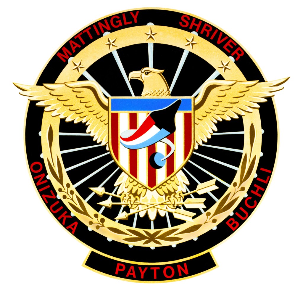 A round black and gold mission patch featuring a gold eagle with a shield of red and white vertical stripes and a blue strip along the top and a black image of a space shuttle flying across it; the names of the five astronauts are written in red letters around the black perimeter of the patch.