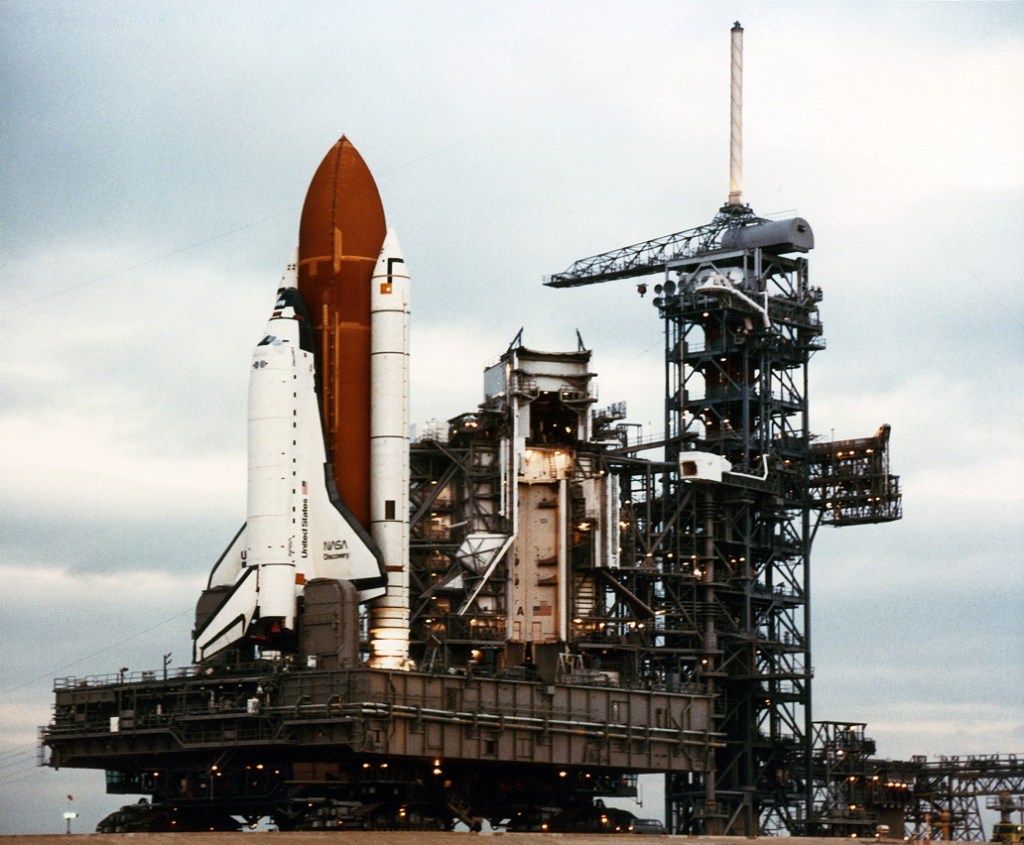 Image of a space shuttle standing vertical on the crawler as it approaches its launch pad during rollout