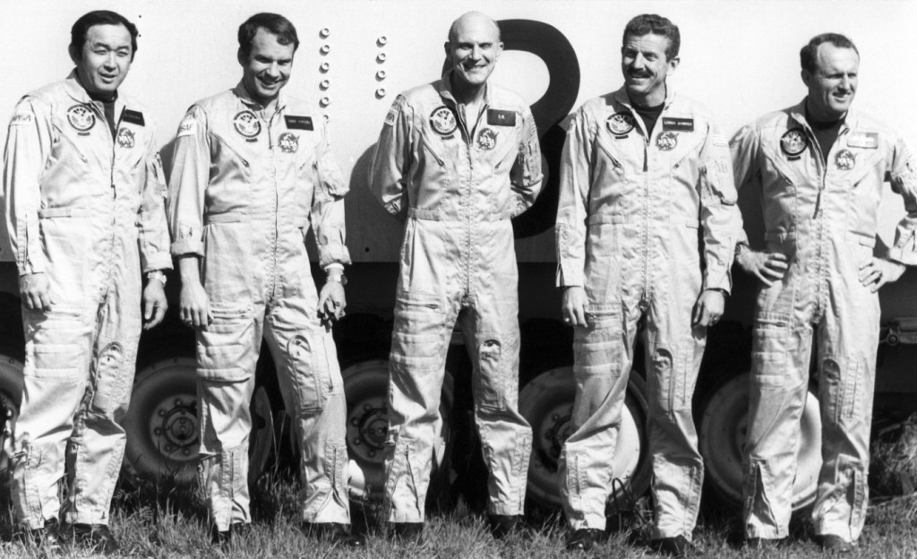 Black and white image of five men wearing flight suits standing side by side.