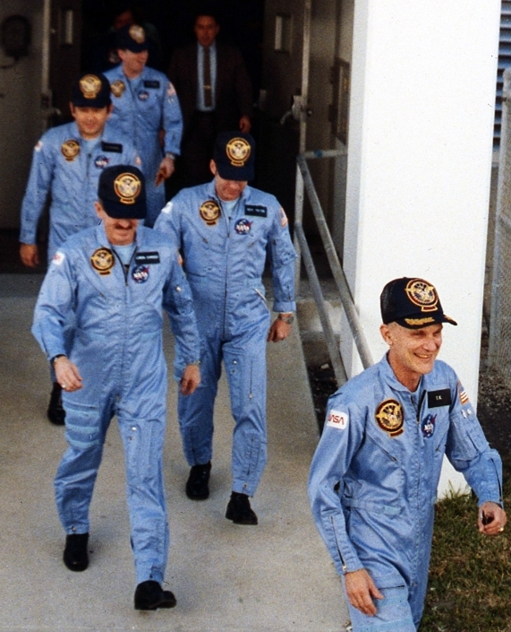 Image of five men wearing blue flight suits and black baseball caps walking out of a building.