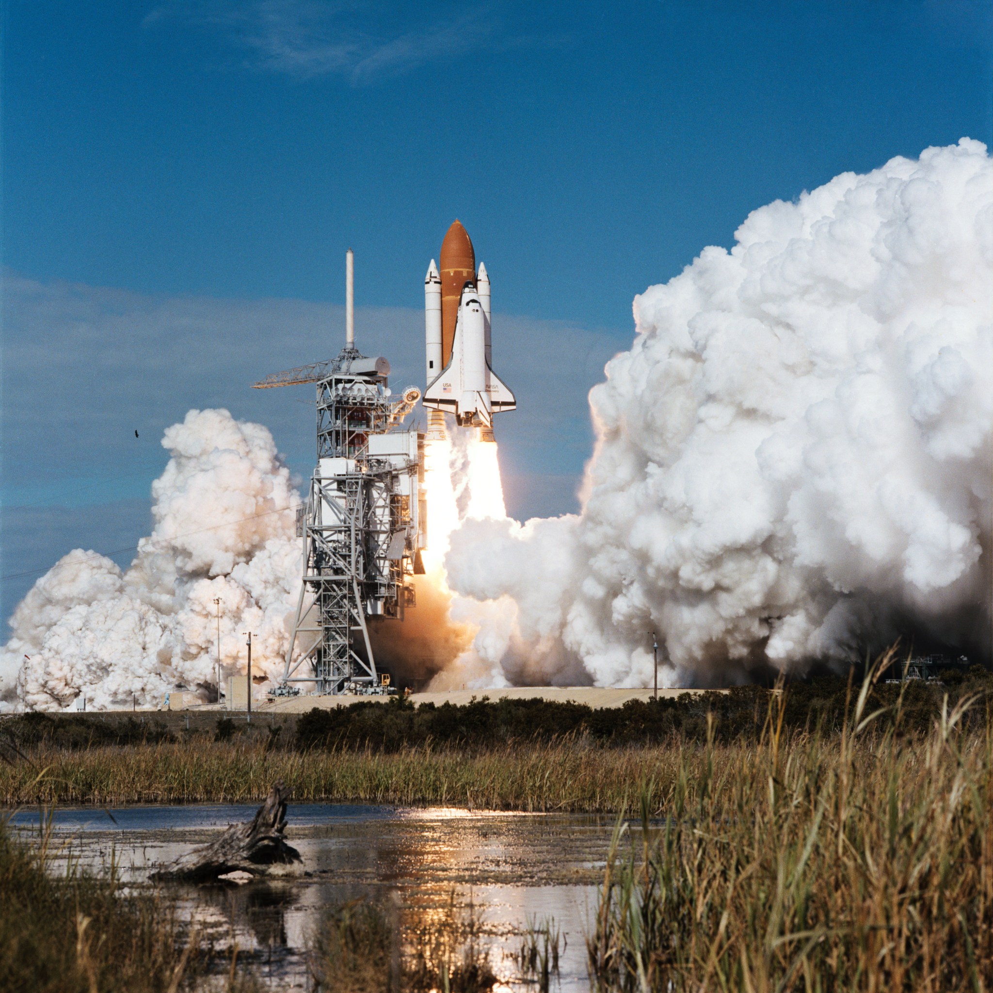 Image of a space shuttle lifting off from its launch pad on a pillar of fire against a blue sky.