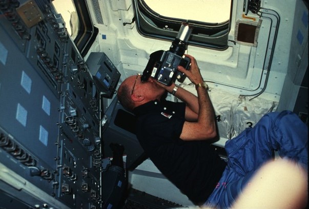 Image of an astronaut holding a camera to a shuttle window while floating in space.